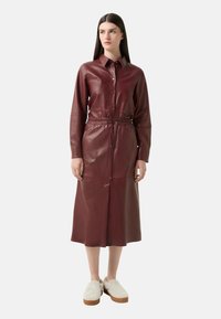Bordeaux leren shirtjurk met een door knopen sluiting, lange mouwen, elastische taille en zijslitsen. Gecombineerd met beige fuzzy instap schoenen.