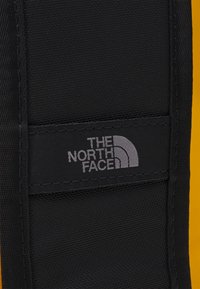 Crni materijal s teksturiranim završetkom ima sivi logo zakrpu s natpisom "THE NORTH FACE" i polukružni dizajn na žutoj pozadini.
