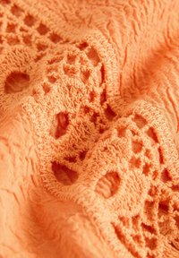 Persikofärgad tyg med texturerad yta och intrikata broderade utskärningsmönster. Mjukt material med en lätt skrynklig yta.
