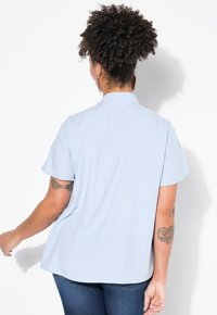 Blusa azzurra a maniche corte con colletto, realizzata in tessuto morbido; orlo curvo e texture liscia visibile sul retro.