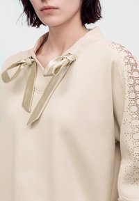 Gros plan d’une personne portant un haut beige avec des nœuds en satin au col et un motif perforé décoratif sur la manche.