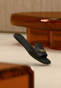 Μαύρο δερμάτινο σαγιονάρι slide με φαρδιά λουρίδα, διακοσμημένο με χρυσή αλυσίδα. Λεπτή σόλα με λεία υφή. Μινιμαλιστικός σχεδιασμός.