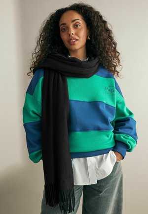 Oversize'owy sweatshirt w zielono-niebieskie paski, z logo z przodu, w połączeniu z czarnym szalikiem. Pod spodem widoczna jest biała koszula.