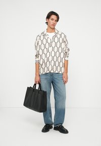 Camicia da uomo a motivi color crema con forme geometriche nere, abbinata a jeans blu chiaro e una borsa a tracolla in pelle nera. Texture liscia, vestibilità casual.