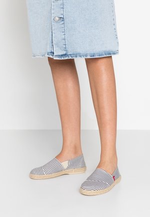 Ben iført blå denim nederdel og slip-on espadrillos med marineblå og hvide striber på lys baggrund.