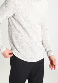 Långärmad ljusgrå topp med ett texturerat prickigt mönster, i kombination med mörka byxor. Tyget verkar mjukt och sitter tajt vid midjan.