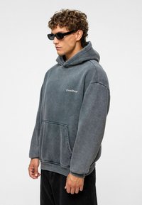Grijze hoodie met een kangoeroe zak en ribbelmanchetten. Bevat de tekst "Create Change" gedrukt op de borst. Gemaakt met hoekige zwarte zonnebril.