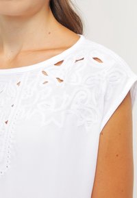 Weißes Blusenshirt mit aufwendiger Stickerei an den Schultern, kurzen Ärmeln und einem runden Halsausschnitt mit ausgeschnittenen Details. Glatte Textur.