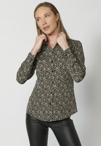 Camicia nera con bottoni, caratterizzata da un motivo floreale in crema e giallo. Maniche lunghe con colletto classico e una silhouette leggermente aderente.