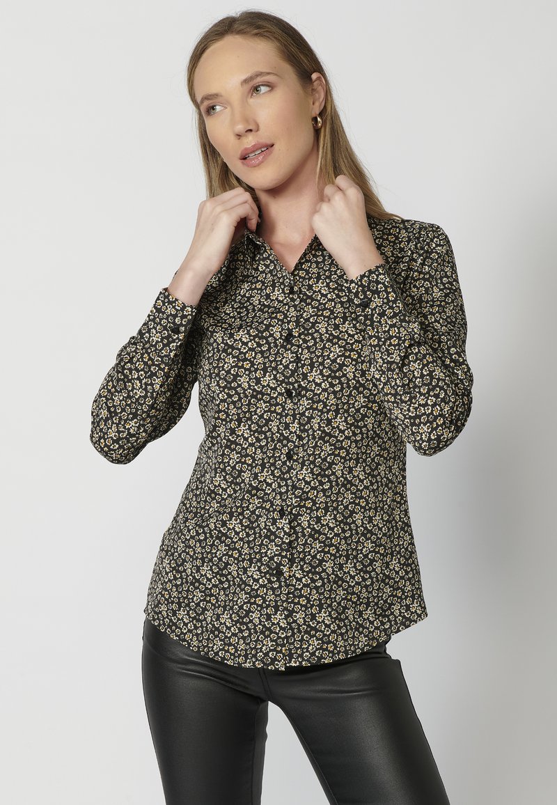 Camicia nera con bottoni, caratterizzata da un motivo floreale in crema e giallo. Maniche lunghe con colletto classico e una silhouette leggermente aderente.