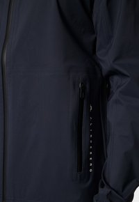 Marinblå vindjacka med en jämn textur, som har en dragkedjeficka och en logotyp på sidan i kontrasterande vitt.