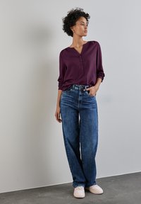 Blusa a maniche lunghe di un profondo colore viola con scollatura a bottoni, abbinata a un paio di jeans blu larghi e sneaker con dettagli rosa.