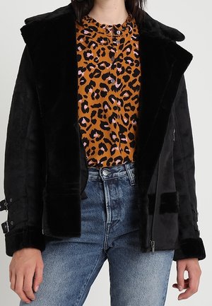 Kobieta ubrana w czarną kurtkę z czarnego futra na sztuczne, nałożoną na brązową i różową bluzkę w panterkę oraz wyblakłe niebieskie dżinsy, dłonie swobodnie opuszczone po bokach.