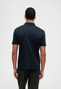 Camisa polo de color navy con cuello, mangas cortas y textura suave, llevada por una persona de espaldas a la cámara frente a un fondo liso.