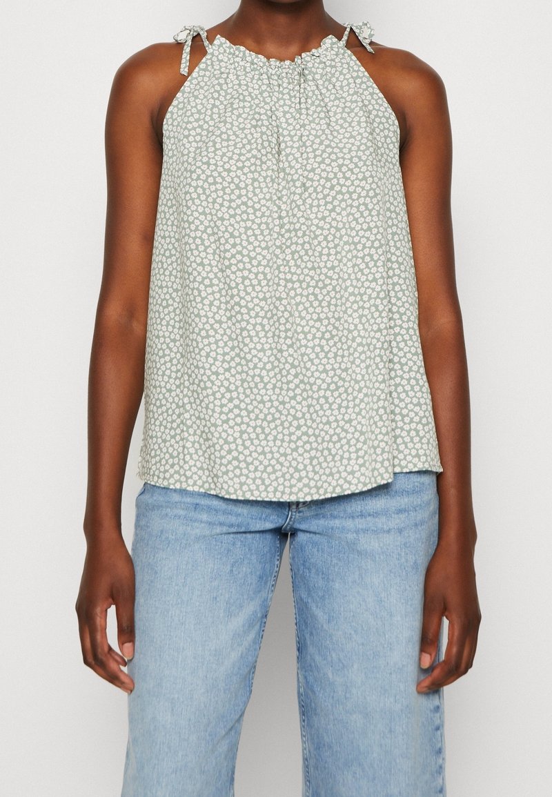 Haut licou vert clair à motif floral, avec bretelles ajustables à nouer et encolure froncée. Associé à un jean en denim bleu clair.
