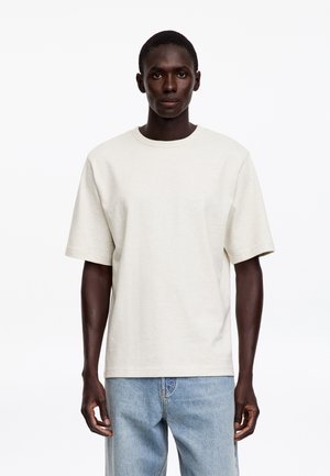 Jeune homme debout face à l’avant portant un t-shirt uni blanc cassé à manches courtes et un jean bleu clair sur un fond blanc.