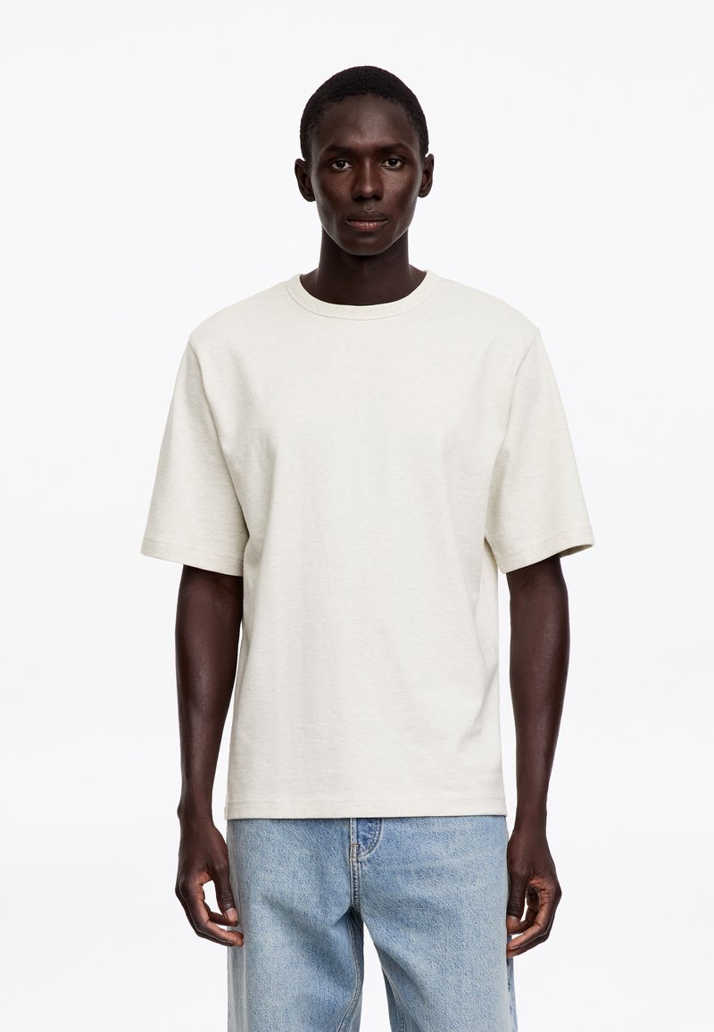 Jeune homme debout face à l’avant portant un t-shirt uni blanc cassé à manches courtes et un jean bleu clair sur un fond blanc.