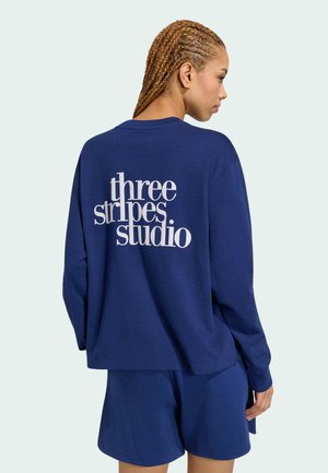 Kvinde med fletninger iført navyblå bluse med lange ærmer og shorts, set bagfra, tekst på blusen lyder "three stripes studio."