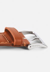 Bracelet de montre en cuir marron avec une boucle en métal argenté poli. Présente des détails de couture et une gravure de marque sur la boucle.