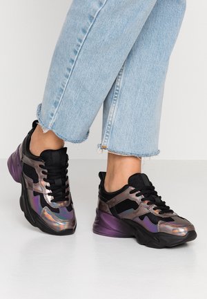 Person wearing light blue cropped jeans and black-purple iridescent chunky sneakers with black laces on a white floor.