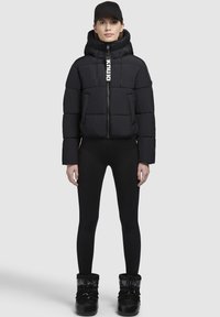 Puffig svart jacka med hög krage och zip-stängning, med en justerbar huva och logodetalj. Buren med svarta leggings och stövlar.