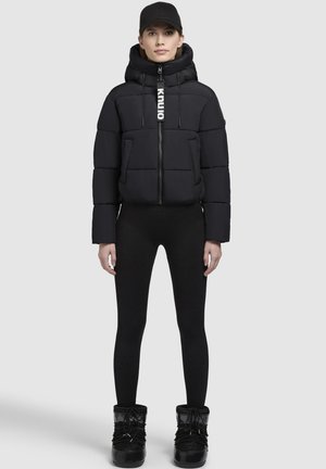 Giacca nera imbottita con colletto alto e chiusura con zip, dotata di cappuccio regolabile e dettaglio con logo. Indossata con leggings neri e stivali.