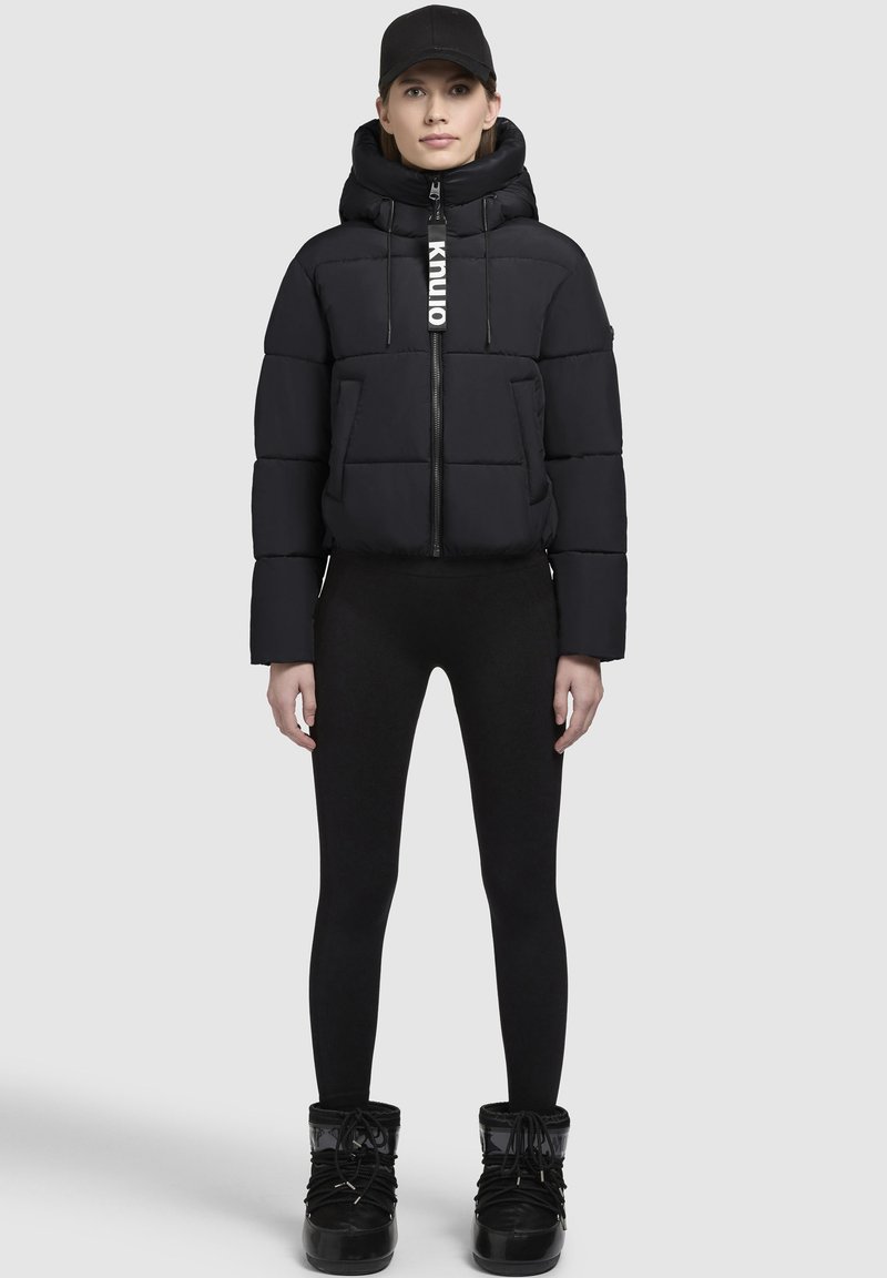 Puffig svart jacka med hög krage och zip-stängning, med en justerbar huva och logodetalj. Buren med svarta leggings och stövlar.