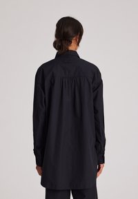 Camicia nera a maniche lunghe con colletto aderente, schiena plissettata e polsini con bottoni. Realizzata in tessuto leggero con una texture liscia.