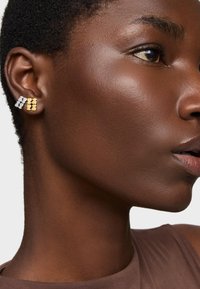 Złote i srebrne geometryczne kolczyki w kształcie studa, metaliczne wykończenie, kontrastujące kształty, prezentowane na ciemnej karnacji skóry na neutralnym tle.