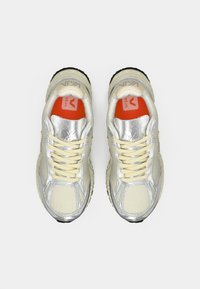 Jasno beżowe sneakersy z srebrnymi akcentami, teksturowana siateczka na górze, beżowe sznurówki i czarna podeszwa. Posiadają pomarańczowy detal logo na wkładce.
