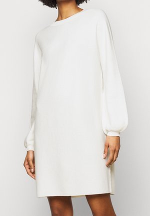 Mujer con un vestido blanco de punto, de manga larga y largo hasta la rodilla, con mangas abullonadas y puños acanalados, sobre un fondo liso.