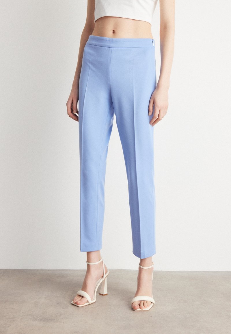 Light blue tailored trousers with a smooth texture, tapered legs, and a flat front. White heeled sandals complement the outfit.
