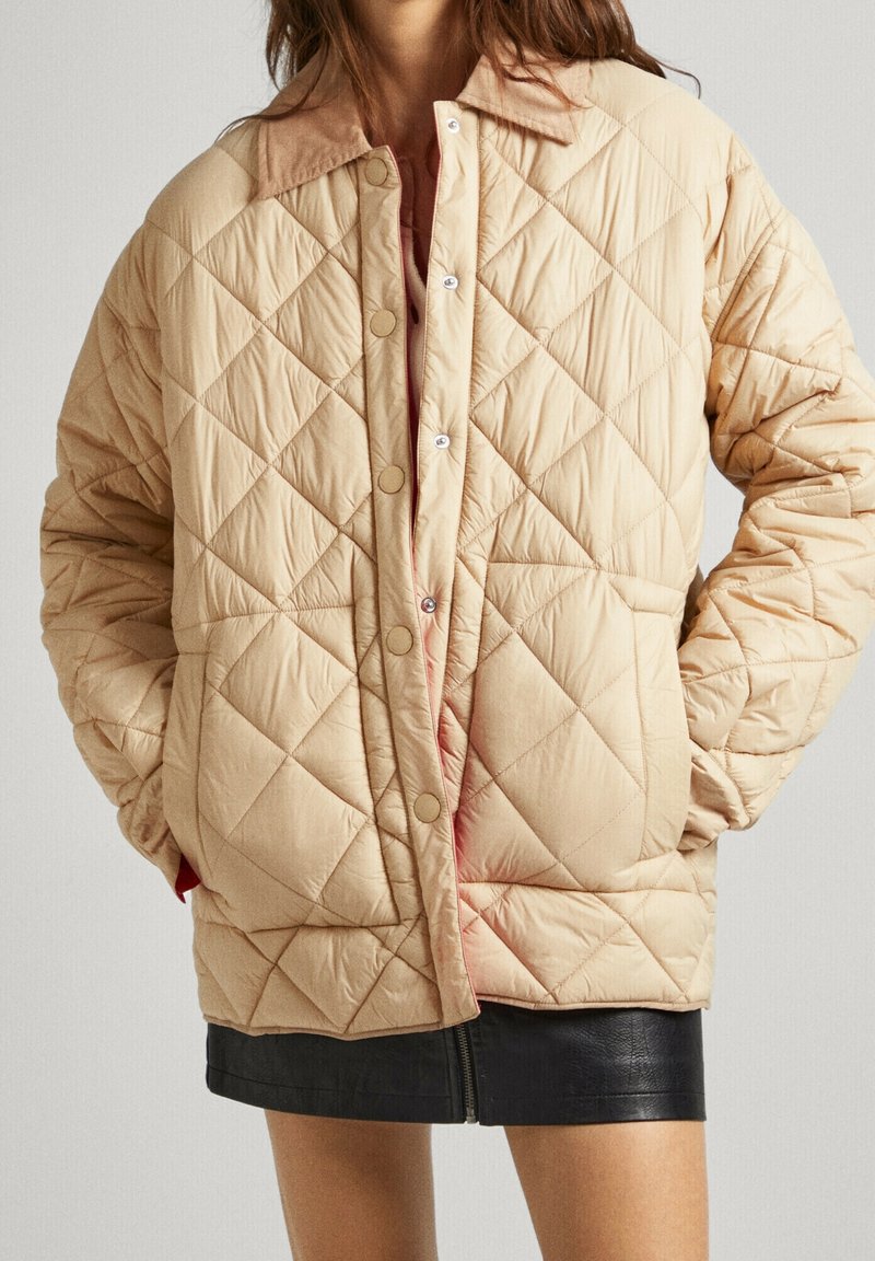 Femme portant une veste matelassée beige avec des boutons-pression, un col en velours côtelé, les mains dans les poches et une mini-jupe en cuir noire.
