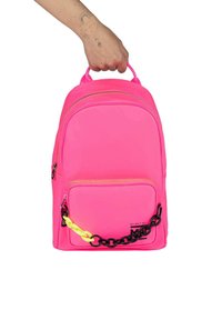 Mochila de nylon rosa con un asa superior, bolsillo frontal con cremallera y una correa de cadena negra adornada con una trenza amarilla. Textura suave y diseño redondeado.