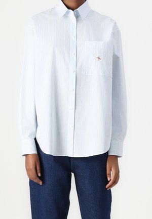 Person wearing a light blue and white striped button-down shirt with a chest pocket and dark blue jeans, shown from neck to hips.