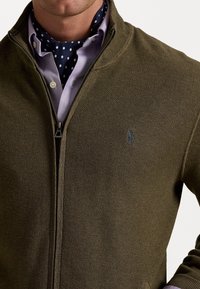 Muž v olivově zeleném svetr s zipem a texturou přes světle fialovou košili, s námořnickou modrou šálou s puntíky kolem krku.