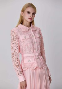 Femme aux longs cheveux blonds portant un haut en dentelle rose avec des boutons en perles et une jupe plissée assortie, se tenant devant un fond uni.