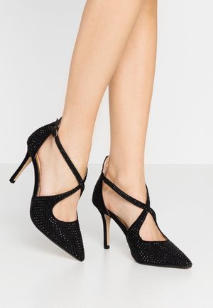 Vrouw met zwarte pumps met spitse neus, enkelbandjes en kleine strass steentjes, staande op een witte achtergrond.