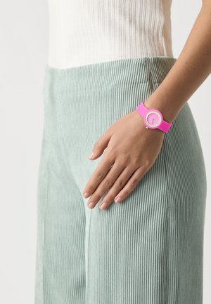 Montre en silicone rose avec un cadran rond, des accents argentés et un bracelet texturé. Portée sur une main sur un fond de tissu vert clair à rayures.
