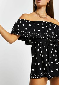 Kvinna som bär en svart off-shoulder klänning med vita prickar, ett guld-halsband och guld-hoop örhängen mot en enfärgad bakgrund.