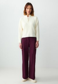 Crème ribbed pullover met een half-zip, gecombineerd met brede bordeauxrode broek en beige sneakers. Soepele textuur, comfortabele pasvorm en contrasterende kleuren.
