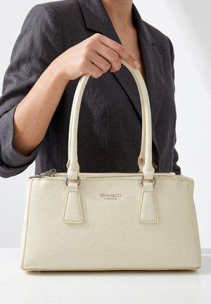Vrouw in een donkere blazer houdt een crèmekleurige Diana & Co Firenze handtas met dubbele handvatten op een witte ondergrond.
