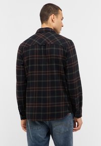 Chemise en flanelle à carreaux sombres avec des rayures marron et vertes, poignets à boutons et ourlet arrondi. Le tissu a une texture douce, idéale pour les tenues décontractées.