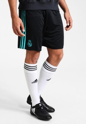 Homme portant un short noir du Real Madrid avec des bandes turquoise, des chaussettes blanches Adidas montant jusqu'aux genoux avec des bandes noires, et des crampons Adidas noirs.