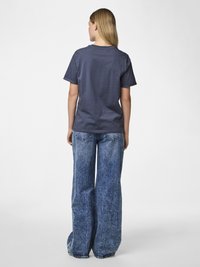 Kobieta ubrana w ciemnoszarą koszulkę i szerokie dżinsy z efektem sprania, stojąca tyłem do aparatu na jednolitym białym tle.