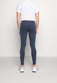 Un homme portant des leggings ajustés bleu foncé et des baskets blanches, se tenant devant un fond gris clair uni, de dos.