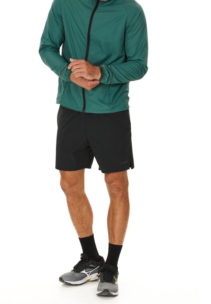 Personne portant une veste verte zippée, un short noir, des chaussettes noires et des chaussures de course grises, debout, les mains jointes à la taille.