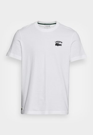 Biała koszulka z krótkim rękawem i okrągłym dekoltem, z małym czarnym logo Lacoste oraz emblematem krokodyla na lewej piersi.