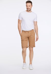 Kahvimpi puuvillashortsit, joissa on tasainen etuosa, käännettävät hihansuut ja taskut, yhdistettynä yksinkertaiseen valkoiseen pyöreäkaulaiseen t-paitaan ja valkoisiin lenkkareihin.