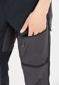 Svarta och grå utomhusbyxor med flera zipfickor, texturerat tyg och förstärkta sömmar för hållbarhet. Hand inskjuten i zipfickan.